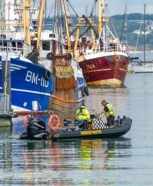 Brixham, UK. 10 August 2021. Two men on the Harbour Master tender boat leave Brixham Harbour with trawlers behind.