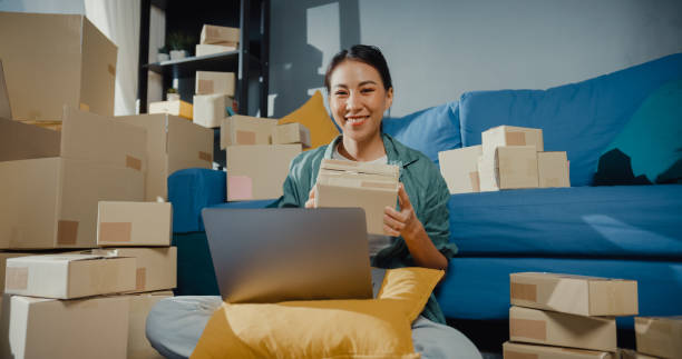 istock Young Asian female seller worker online store using laptop preparing parcel for shipment to client at home. 1363123152