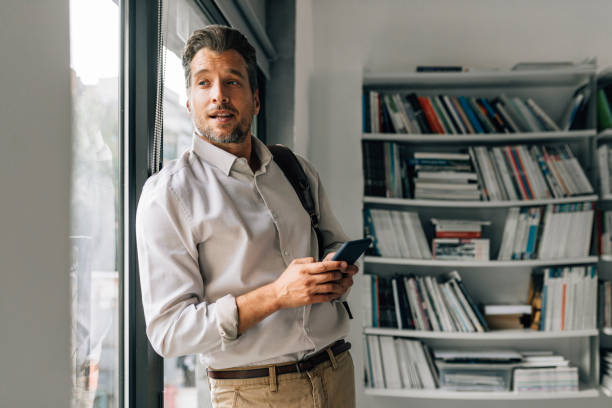 istock Handsome Teacher Standing in the Library and Using his Mobile Phone 1363123442