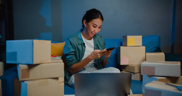istock Young Asian female seller worker online store using phone preparing parcel for shipment to client at home at night. 1363123609