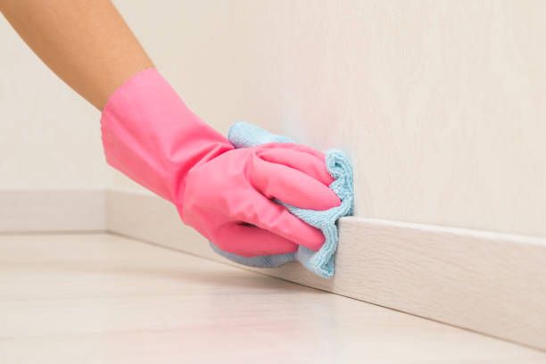Young adult woman hand in pink rubber protective glove using blue dry rag and wiping light wooden baseboard surface in room at home. Closeup. stock photo