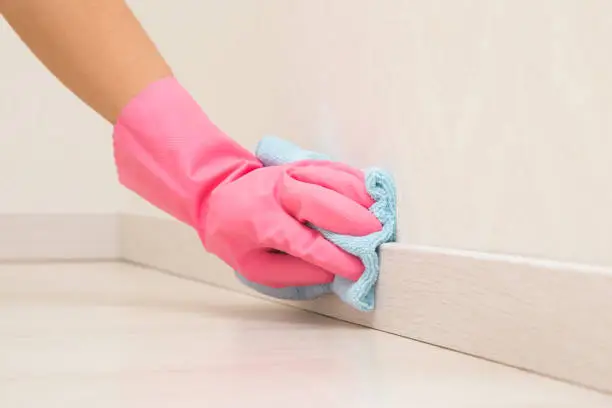 Photo of Young adult woman hand in pink rubber protective glove using blue dry rag and wiping light wooden baseboard surface in room at home. Closeup.