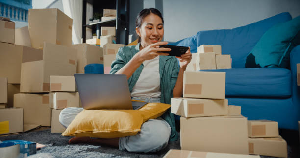 istock Young Asian female seller worker online store using mobile app on smartphone checking parcel box for shipment to client at home. 1363126132