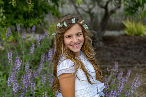 Brown-haired girl smells flower and poses near pinkish flowers on green bush lit by sunlight. Schoolgirl with long braids enjoys posing and taking picture