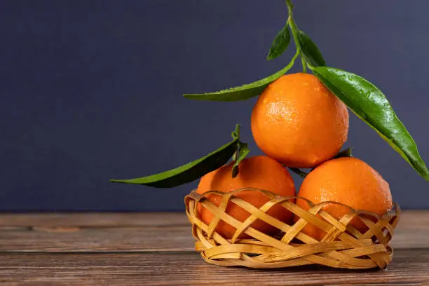 Three fresh ripe mandarin orange tangerine with green leaves in a woven wicker basket isolated on a wooden table against a blue background. Copy space for text. Juicy orange-colored fruit. A closeup.