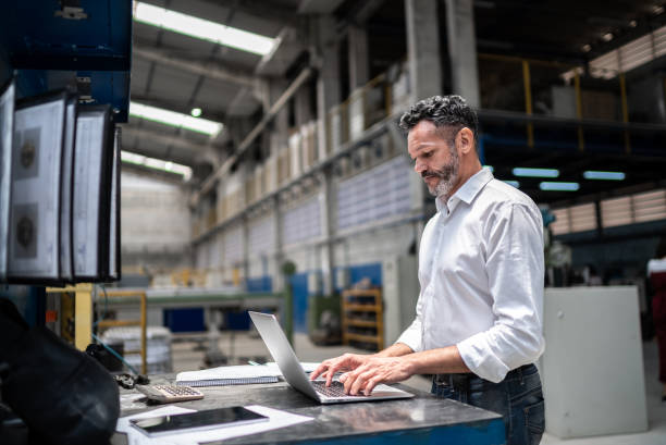 mature businessman using laptop in a factory - indústria imagens e fotografias de stock