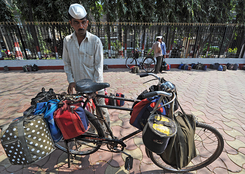 Tiffin wallah or servant delivers tiffin boxes with employer's lunch slung on his bicycle, Mahatma Ghandi Road, Mumbai, Maharashtra, India. Tiffin is a British tradition carried through to today and many of the lunches have already been delivered and hung on the railings behind the cyclist. Mumbai, Maharashtra, India. Mumbai is the second most populous city in India built over seven islands and attracts poor people from all over India who beg and scrape a subsistence living in the face of great wealth from the indigenous Indian people and global tourists