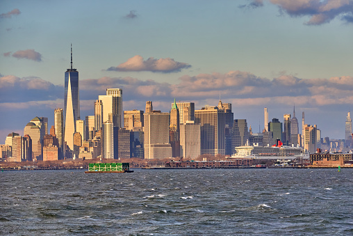 New York, NY - December 22, 2021: A golden light shines on the skyline of Lower Manhattan and the Queen Mary 2 Ocean Liner, docked at the The Brooklyn Cruise Terminal in Red Hook, NYC. As seen across the water of New York Harbor.