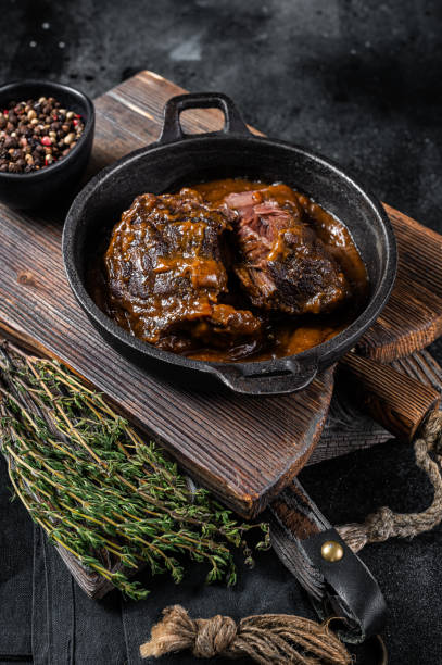 istock Stewed beef cheeks with brown red wine sauce in skillet. Black background. Top view 1363145254