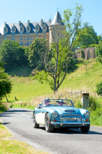 Austin Healey 3000 classic convertible sportscar touring with driver and passenger on a country road in front of the majestic Chateau de Rochechouart, Haute-Vienne, France. Classic and historic architecture complimented by this classic automotive history with this old British sportscar, cruising on a sunny day in the French countryside