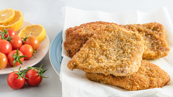Crispy pan fried breaded pork chops served with lemon and fresh tomatoes close up on a plate on light grey background