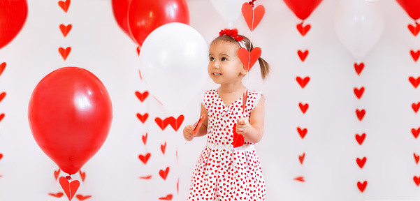 Little baby girl with balloons and a heart-shaped lollipop looks thoughtfully aside at copy space. Decorations on a white studio background for Valentine's Day.