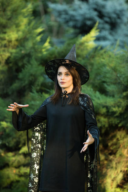 Portrait of an attractive young woman in a black dress with a cape on a background of green forest. Portrait of an attractive young woman in a black dress with a cape on a background of green forest. The girl is wearing a black pointed hat with wide brim. A girl with dark hair and brown green eyes. n11 stock pictures, royalty-free photos & images