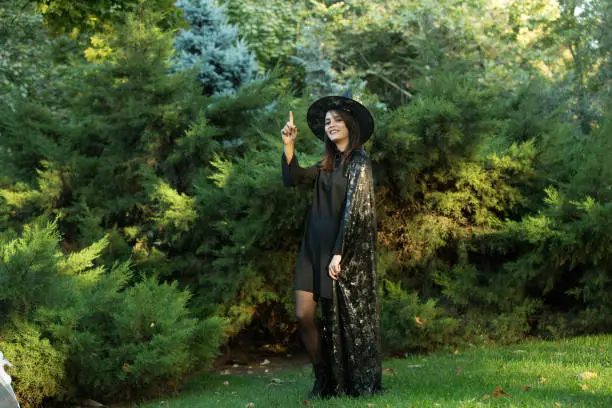 Portrait of an attractive young woman in a black dress with a cape on a background of green forest. The girl is wearing a black pointed hat with wide brim. A girl with dark hair and brown green eyes.