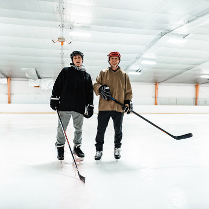 Mature Caucasian man and his young adult son posing for photo at the hockey practice. Both wearing full hockey gear. Interior of Hockey rink indoors.