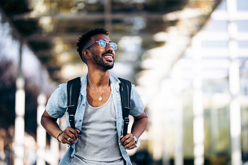 Happy and smiling black man with glasses and backpack walking down the street. Taking the handles of the backpack with his hands.