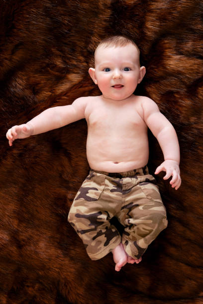 un drôle de petit garçon heureux est allongé sur un tapis de fourrure brune dans un uniforme militaire. enfant souriant avec un ventre nu dans des vêtements kaki, vue de dessus - fake fur white happiness beginnings photos et images de collection