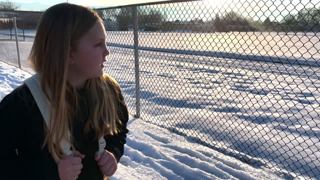 8-9 year old girl walking home from school in the snow along chainlink fence