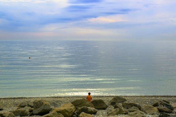 A beach on the Black Sea coast. A man among boulders looks at the sea under a blue cloudy sky. Adler, Sochi, Russia, 2021
