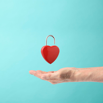 Unlocking red heart shape padlock floating in mid- air above hand, against light blue background.