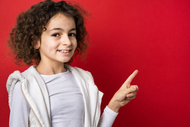 one small caucasian girl ten years old with curly hair front view portrait close up standing in front of red background looking to the camera pointing finger to copy space smiling happy and joy - 10 11 years child human face female imagens e fotografias de stock