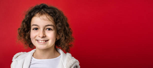 곱슬 머리 앞보기 초상화와 함께 10 살 된 작은 백인 소녀 한 명이 행복하고 즐거운 복사 공간을 미소 짓는 카메라를 바라보며 빨간색 배경 앞에 서 있습니다. - 10 11 years little girls child serious 뉴스 사진 이미지