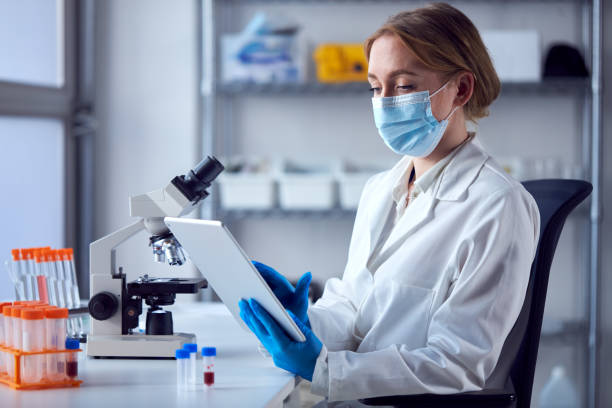 istock Female Lab Worker Wearing Lab Coat Working On Covid-19 Vaccine With Digital Tablet And Microscope 1363236452