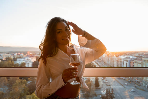 Beautiful young brunette with long curly hair standing on rooftop with glass of wine. She looks happy and confident. Cityscape and sunset are in background. Brunette with long curly hair standing next to the window. She is holding glass of wine. She looks happy and confident. Cityscape and sunset are in background. golden hour wine stock pictures, royalty-free photos & images