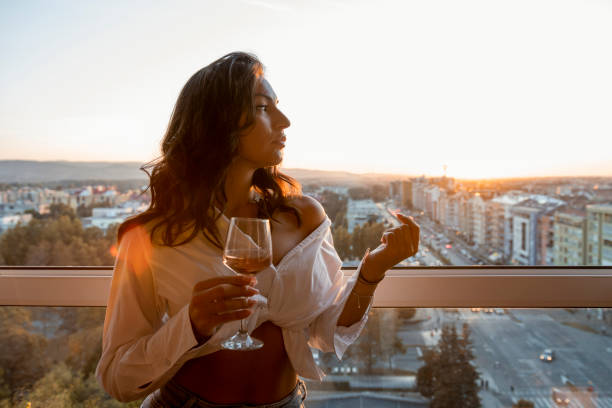 Beautiful brunette with long curly hair holding glass of wine. She is standing next to the window and watching sunset. Urban background. Young brunette with long curly hair standing next to window and holding glass of wine. Sunset and urban town visible in background. after work photos stock pictures, royalty-free photos & images