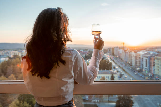 Brunette enjoying sunset. She is holding glass of wine next to window. Cityscape in the background. Woman with long, curly, brown hair standing next to the window with glass of wine. She is watching sunset. Cityscape in the background. golden hour wine stock pictures, royalty-free photos & images