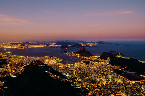 Brazil, Rio de Janeiro. Meeting the sunset in orange-violet shades. Top view of the city, lanterns, ocean, hills, light from lanterns, buildings and cars.
