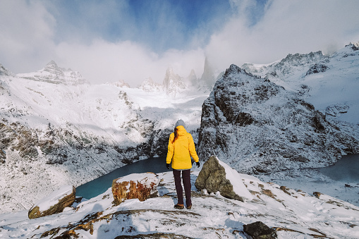 Girl on mountain peak with snow and lake looking at beautiful mountain valley in cloud in swinter Landscape with sporty young woman, cloudy hills, sky. Travel and tourism. Hiking
