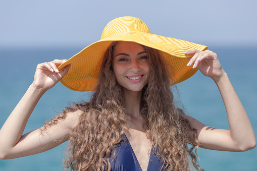 Waist-up portrait of pretty girl against sea. Curly young woman in yellow board brim hat poses with arms raised and looks at camera smiling