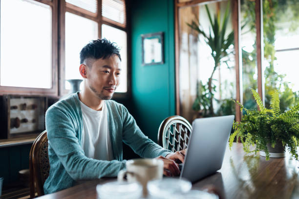 Smiling young Asian man sitting at dining room table at home, shopping online with laptop. Convenient and safe online payment concept. Technology makes life much easier stock photo