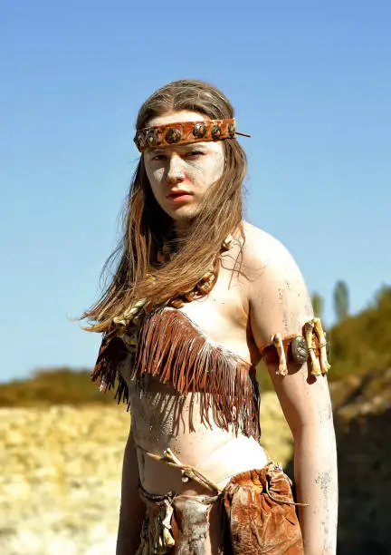 A young woman is dressed as Neanderthal warrior. 
Her body and face is covered with mud and dirt.
She is seen posing in a stone quarry.