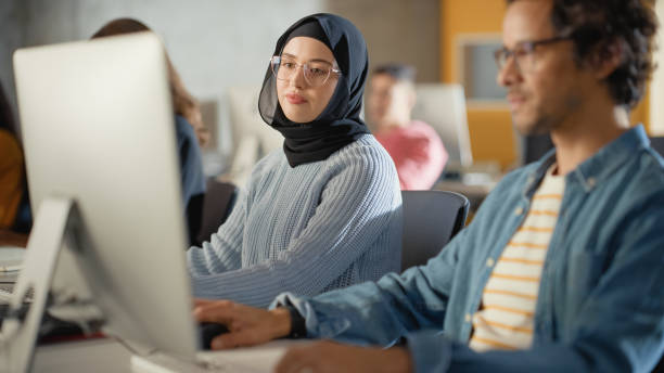 istock Curious Female Muslim Student Wearing a Hijab, Studying in Modern University with Diverse Multiethnic Classmates. College Scholars Work in College Room, Learning IT, Programming or Computer Science. 1363276344