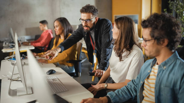 il docente aiuta lo studioso con il progetto, consigliando il loro lavoro. insegnante che dà lezioni a diversi gruppi multietnici di studenti di sesso femminile e maschile in una stanza del college, insegnando nuove abilità accademiche su un computer. - lettore foto e immagini stock