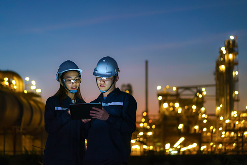 Ingenieros asiáticos, hombres y mujeres están revisando el mantenimiento de la fábrica de la refinería de petróleo por la noche a través de tabletas digitales