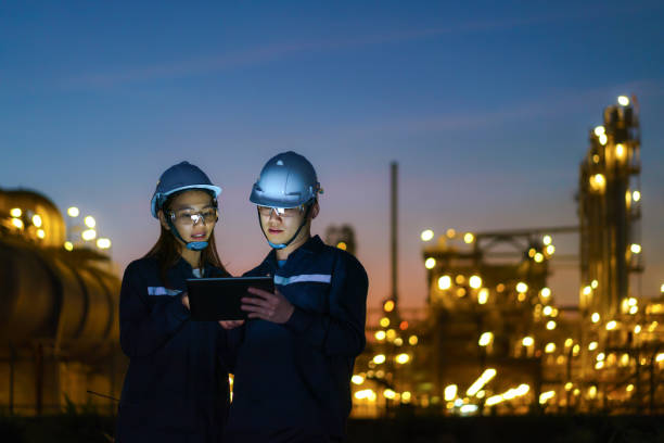 des ingénieurs asiatiques, hommes et femmes, vérifient la maintenance de l’usine de raffinerie de pétrole la nuit via des tablettes numériques. r - industrie du pétrole photos et images de collection