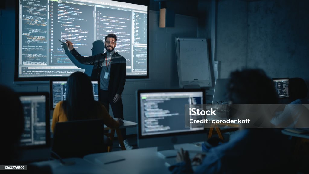 Teacher Giving Computer Science Lecture to Diverse Multiethnic Group of Female and Male Students in Dark College Room. Projecting Slideshow with Programming Code. Explaining Information Technology. Technology Stock Photo