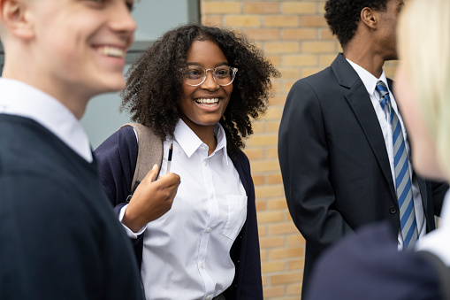 Candid view of cheerful multiracial schoolgirl in uniform, eyeglasses, and backpack standing outdoors with friends, talking and laughing.