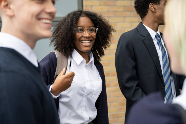 Male and female teenagers interacting between classes Candid view of cheerful multiracial schoolgirl in uniform, eyeglasses, and backpack standing outdoors with friends, talking and laughing. english spoken stock pictures, royalty-free photos & images