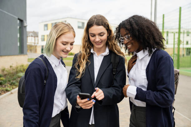 Teenage schoolgirls checking social media on smart phone Waist-up front view of friends in uniforms standing outdoors on secondary campus and smiling as they surf mobile device between classes. teenagers only teenager multi ethnic group student stock pictures, royalty-free photos & images