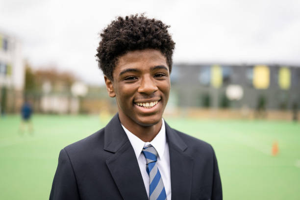 Campus portrait of cheerful Black schoolboy in uniform Head and shoulder front view of 15 year old student in shirt, necktie, and navy blue blazer standing outdoors on sports field and smiling at camera. schoolyard photos stock pictures, royalty-free photos & images