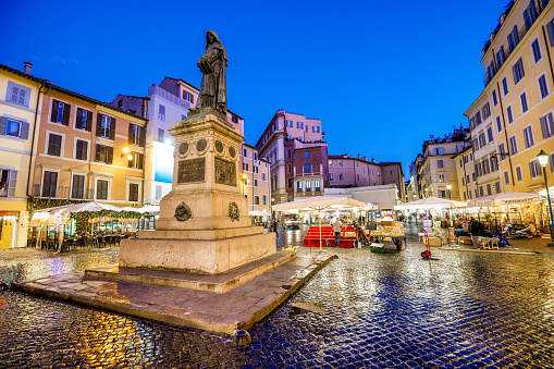 A suggestive evening view of the picturesque Campo de Fiori square, located in the historic and baroque heart of Rome, between Piazza Farnese and Piazza Navona. In the center, the bronze statue of the religious and philosopher Giordano Bruno, created by the sculptor Ettore Ferrari in 1889, erected in the place where Bruno was condemned and burned at the stake by the Holy Inquisition in 1600. The Campo de Fiori district is an area of the Eternal City much loved and visited by tourists and residents for the presence of ancient churches and noble palaces and for the countless artistic and cultural treasures of the Renaissance and Baroque era. But Campo de Fiori is also famous for the characteristic flower, fruit and vegetable market that occupies the square every day and for the presence of numerous trendy restaurants and pubs. Around the square, in the ancient Parione and Regola districts, you can explore dozens of characteristic alleys, where you can discover the essence and soul of Roman life. In 1980 the historic center of Rome was declared a World Heritage Site by Unesco. Super wide angle image in high definition format.