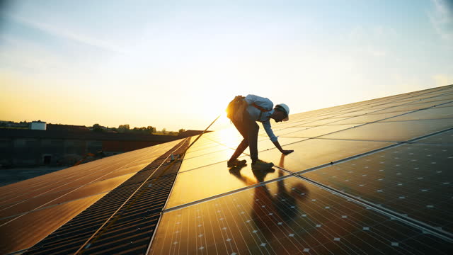 Maintenance assistance technical worker in uniform is checking an operation and efficiency performance of photovoltaic solar panels on roof at sunset. Front view
