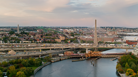 Drone shot of the Charles River in Boston at sunset, looking towards the Leonard P. Zakim Bunker Hill Memorial Bridge. There is heavy freeway traffic on the bridge, and a train is headed out of North Station on the drawbridge below. \n \nAuthorization was obtained from the FAA for this operation in restricted airspace.