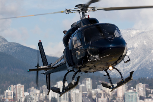 Dark blue helicopter on approach for landing with buildings and mountains in the background.