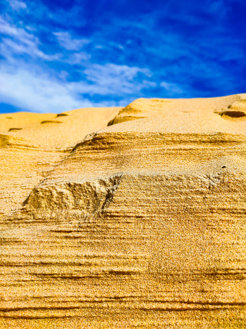 A sandy mountain against a blue sky with clouds - photo of a sand dune against a blue sky with white clouds. Small grains of sand. Dune background. The wind blew away the sand. Clouds in the sky. A mountain of sand and the sky.