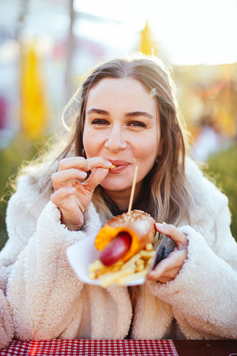 Happy woman eating a hot dog. Woman with sandwich outdoors. Close-up of young blonde woman in red cap and eco fur coat is eating out sandwich at sandwich bar at street fair.